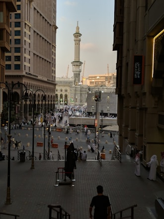 A bustling cityscape featuring a large, ornate mosque tower with detailed architecture, surrounded by high-rise buildings. The street is filled with people, many in traditional attire, suggesting a busy urban environment.