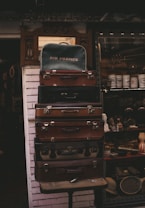 A stack of vintage suitcases is displayed against a brick wall. The top suitcase is labeled with 'Air France'. Behind the suitcases, a shop window showcases various antiques, including jars, figurines, and a violin.