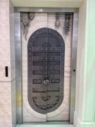 A metal elevator door designed to resemble a medieval vault door with rivets and decorative stone-like elements. The door features a symmetrical pattern and is set within a tiled wall with a small control panel displaying the floor number 711.