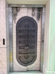 A metal elevator door designed to resemble a medieval vault door with rivets and decorative stone-like elements. The door features a symmetrical pattern and is set within a tiled wall with a small control panel displaying the floor number 711.
