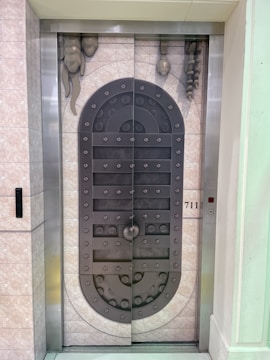 A metal elevator door designed to resemble a medieval vault door with rivets and decorative stone-like elements. The door features a symmetrical pattern and is set within a tiled wall with a small control panel displaying the floor number 711.