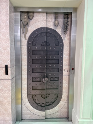 A metal elevator door designed to resemble a medieval vault door with rivets and decorative stone-like elements. The door features a symmetrical pattern and is set within a tiled wall with a small control panel displaying the floor number 711.