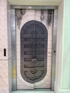 A metal elevator door designed to resemble a medieval vault door with rivets and decorative stone-like elements. The door features a symmetrical pattern and is set within a tiled wall with a small control panel displaying the floor number 711.
