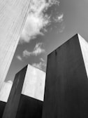 Large concrete slabs rise into the sky, creating a geometric pattern against a backdrop of scattered clouds. The arrangement of the slabs suggests a modern architectural structure, with sharp angles and a minimalistic design.