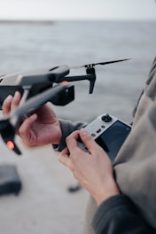 Hands holding a drone and its remote controller near a body of water.