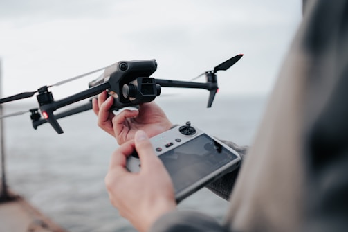 A person operates a drone using a remote controller. The drone is in focus with visible rotors and camera, and is being prepared for flight near a body of water.