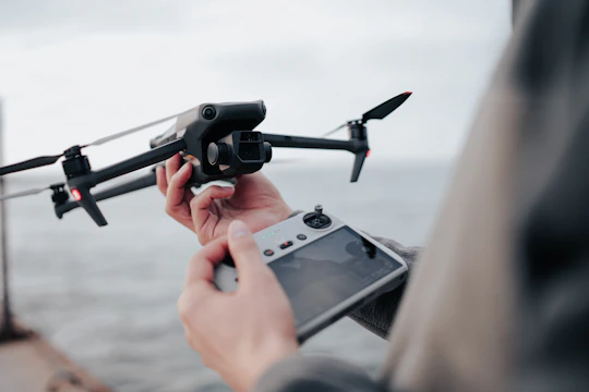 A person operates a drone using a remote controller. The drone is in focus with visible rotors and camera, and is being prepared for flight near a body of water.