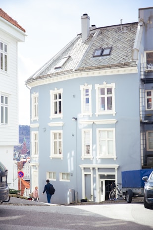 A light blue European-style building on a sloped street with white trim around its windows and doors. The roof is slightly angled with a few skylights. A man with a backpack and a child in a pink outfit are walking past. Bicycles and a vehicle covered with a blue tarp are parked nearby, and a 'no parking' sign is visible. In the background, there are more buildings and a glimpse of a hilly landscape.