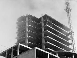 A black and white image of a multi-story building under construction with scaffolding and a large crane visible. The structure has multiple levels with visible concrete slabs and columns. The atmosphere appears foggy or dusty, obscuring some details.