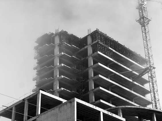 A black and white image of a multi-story building under construction with scaffolding and a large crane visible. The structure has multiple levels with visible concrete slabs and columns. The atmosphere appears foggy or dusty, obscuring some details.