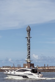 A modern yacht travels across a body of water with a tall, intricately designed sculpture standing in the background. The sky is clear with scattered clouds, creating a serene setting. The sculpture is mounted on a square base, surrounded by a stone barrier, emphasizing a blend of natural and artistic elements.