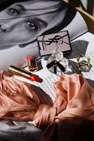 A glossy magazine page with a monochrome portrait partially visible under an arrangement of beauty products. These include a branded perfume bottle with a black ribbon, a closed compact with a logo, and an open lipstick with a red shade. Two textured gold rings and a peach silk fabric with floral patterns are neatly displayed. The soft, diffused lighting creates gentle shadows, adding a touch of elegance to the composition.
