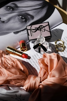 A glossy magazine page with a monochrome portrait partially visible under an arrangement of beauty products. These include a branded perfume bottle with a black ribbon, a closed compact with a logo, and an open lipstick with a red shade. Two textured gold rings and a peach silk fabric with floral patterns are neatly displayed. The soft, diffused lighting creates gentle shadows, adding a touch of elegance to the composition.