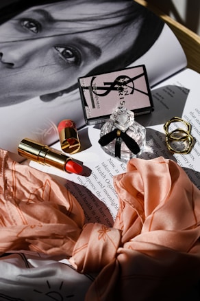 A glossy magazine page with a monochrome portrait partially visible under an arrangement of beauty products. These include a branded perfume bottle with a black ribbon, a closed compact with a logo, and an open lipstick with a red shade. Two textured gold rings and a peach silk fabric with floral patterns are neatly displayed. The soft, diffused lighting creates gentle shadows, adding a touch of elegance to the composition.