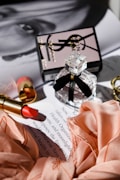 A flat lay composition featuring a square glass perfume bottle with a black ribbon, a gold tube of red lipstick, a silk scarf in a peach color, and a product box with a logo. These items are placed on top of a magazine page with partial text visible.