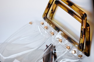 A transparent plastic handbag with a brown tortoiseshell-patterned handle and gold rivets along the top edge. The clear material creates a sleek and modern look, reflecting light subtly. The design combines minimalist and contemporary styles.