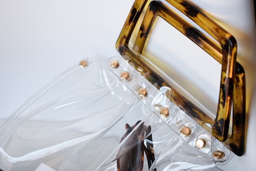 A transparent plastic handbag with a brown tortoiseshell-patterned handle and gold rivets along the top edge. The clear material creates a sleek and modern look, reflecting light subtly. The design combines minimalist and contemporary styles.