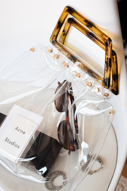 A clear, transparent bag with tortoiseshell handles and gold studs contains a pair of sunglasses, a black case, and an Acne Studios label. The bag rests on a white surface along with two pieces of jewelry visible near the bottom.