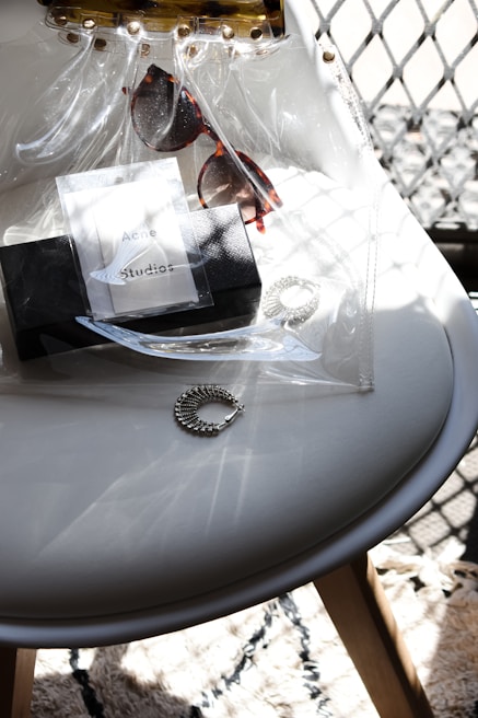 A transparent bag with gold studs contains a pair of tortoiseshell sunglasses, a branded black box, and a silver bracelet, all placed on a white chair. Sunlight casts intricate shadows and highlights on the seat and objects, creating a pattern on the textured rug below.