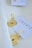 A pair of intricately designed gold earrings are placed on a cream-colored fabric. Each earring has a detailed circular pattern with a small dark blue stone at the center. A white card nearby has the word 'LIBERTE' printed on it in black lettering.