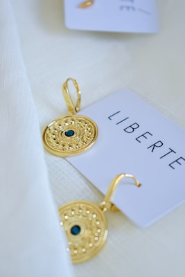 A pair of intricately designed gold earrings are placed on a cream-colored fabric. Each earring has a detailed circular pattern with a small dark blue stone at the center. A white card nearby has the word 'LIBERTE' printed on it in black lettering.