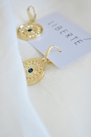 Two gold earrings with intricate designs rest on a textured white fabric. Each earring features a dark central stone and a detailed circular pattern. A card with the word 'LIBERTE' in bold, minimalist font is partially visible.