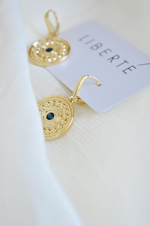Two gold earrings with intricate designs rest on a textured white fabric. Each earring features a dark central stone and a detailed circular pattern. A card with the word 'LIBERTE' in bold, minimalist font is partially visible.