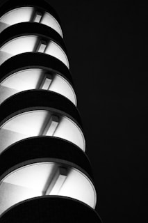 A black and white photograph of a modern, cylindrical building or structure with several levels. Each level is illuminated from within, creating a striking contrast against the dark background. The design features repetitive, curved elements with strong lines.
