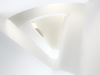 A minimalistic architectural structure featuring smooth, curved lines and a predominantly white aesthetic. The image has an abstract quality with light and shadows creating a sense of depth and dimension. An air vent is visible, integrated seamlessly into the design.