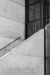 A modern architectural structure featuring a minimalist style with large concrete walls intersected by a series of clean, sharp lines. The lines form part of a staircase with a railing and are contrasted by large glass windows creating a geometric pattern. The overall composition is symmetrical and highlights the interplay between concrete and glass materials.