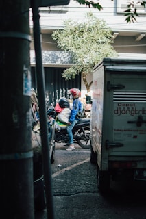 A narrow street scene featuring a motorcyclist wearing a helmet and carrying a passenger. The motorbike is positioned between a parked car and a delivery truck. A leafy tree is visible in the background, adding to the urban setting.