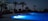 A serene swimming pool is lit up with blue lights during night time, surrounded by palm trees and a residential building in the background. The sky is dark, suggesting it's late evening or night. Some lights from buildings in the distance can be seen on the horizon.