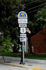 A street sign indicating the beginning of the Florida Keys Scenic Highway, displaying a 'Begin North' notice with a symbolic line marking Mile 0. The background features lush green trees and a brick building.