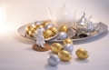 A silver platter holds a collection of shiny, metallic gold and silver eggs. In the background, a white teapot and an ornate silver teapot are visible. In the foreground, a small white angel figurine sits on a wooden base, adding a whimsical touch to the scene.