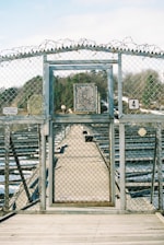 A metal gate with a chain-link fence features barbed wire on top, situated on a wooden deck. Beyond the gate, a long, narrow pathway extends into the distance with rows of metallic structures on either side. A weathered sign with graffiti is mounted on the gate, and a number 4 sign is visible. Trees and a partly cloudy sky can be seen in the background.