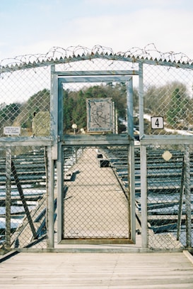 A metal gate with a chain-link fence features barbed wire on top, situated on a wooden deck. Beyond the gate, a long, narrow pathway extends into the distance with rows of metallic structures on either side. A weathered sign with graffiti is mounted on the gate, and a number 4 sign is visible. Trees and a partly cloudy sky can be seen in the background.