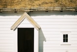 A white wooden building with horizontal planks and a small, triangular-roofed entrance. Above the entrance is a roof covered with yellowed grass, suggesting a rustic, eco-friendly design. A small, square window with a simple frame is visible on the right side.