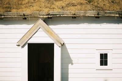 A white wooden building with horizontal planks and a small, triangular-roofed entrance. Above the entrance is a roof covered with yellowed grass, suggesting a rustic, eco-friendly design. A small, square window with a simple frame is visible on the right side.
