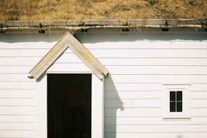 A white wooden building with horizontal planks and a small, triangular-roofed entrance. Above the entrance is a roof covered with yellowed grass, suggesting a rustic, eco-friendly design. A small, square window with a simple frame is visible on the right side.