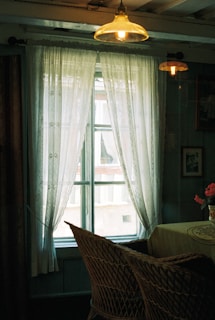 A cozy interior featuring a window framed by sheer lace curtains allowing soft light to filter through. A wicker chair and a table with a floral centerpiece suggest a rustic, vintage atmosphere. The warm glow of hanging pendant lights enhances the inviting ambiance.