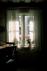 A softly lit room with a wooden table and chairs positioned by a window. Light streams through lace curtains, illuminating a potted plant with red flowers on the windowsill. The room has a vintage feel, with patterned wallpaper and a rustic ceiling beam.