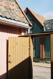 Two wooden houses with traditional, rustic architecture are pictured. The house on the left has pinkish-colored wood panels and a weathered brown door slightly ajar, while the house on the right has greenish-blue wood panels with red trims around the windows. Both houses have sloped, tiled roofs showing signs of age and moss growth. The scene is sunlit, casting strong shadows.