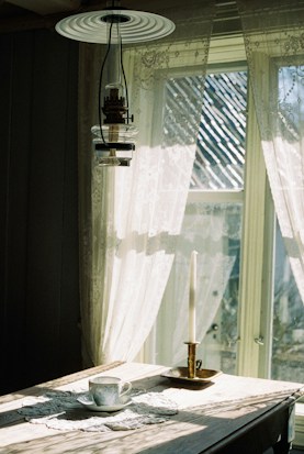 A rustic wooden table adorned with a delicate doily, a fine china teacup and saucer, and an elegant single candle in a brass holder. Sunlight streams through sheer lace curtains, casting intricate patterns of light and shadow across the table.