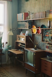 A vintage living room setup with mid-century furniture, including a wooden bookshelf filled with various books and decorative objects. On the shelves, there are colorful plates and framed photographs. A retro television set is prominently featured, alongside an old-style desk with potted plants on the side. The room is well-lit from natural light streaming through a window with sheer curtains, and the walls are decorated with patterned wallpaper.