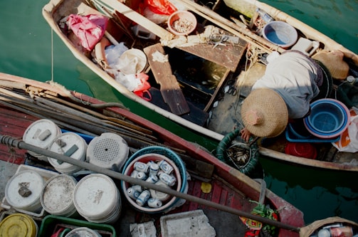 A person wearing a straw hat is working on a small boat surrounded by various containers, baskets, and tools. The scene is situated on water with another boat nearby that has several white buckets and cans onboard.