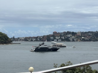 A luxury yacht is sailing on a body of water, with a background of residential buildings and green hills. The scene is partly cloudy, and there are a few smaller boats near the yacht.