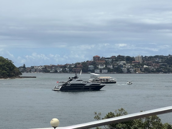 A luxury yacht is sailing on a body of water, with a background of residential buildings and green hills. The scene is partly cloudy, and there are a few smaller boats near the yacht.