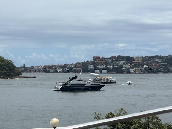 A luxury yacht is sailing on a body of water, with a background of residential buildings and green hills. The scene is partly cloudy, and there are a few smaller boats near the yacht.