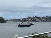 A luxury yacht is sailing on a body of water, with a background of residential buildings and green hills. The scene is partly cloudy, and there are a few smaller boats near the yacht.