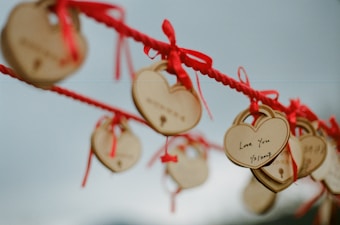 Wooden heart-shaped locks with inscriptions hang on a red rope. The locks are tied with red ribbons, and one is prominently visible with the words 'Love You' and a date written on it. The background is softly blurred, emphasizing the objects and creating a sentimental atmosphere.